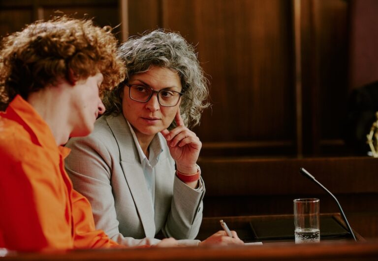 A defense attorney in glasses is consulting with a client in an orange shirt discussing criminal defense strategies during courtroom preparation in Tennessee.