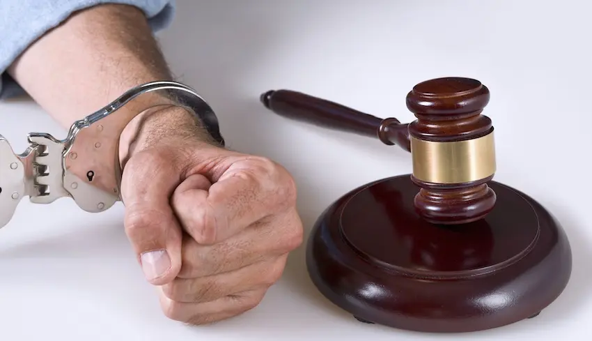 A man's handcuffed fists rest on a table beside a wooden judge's gavel on its sound block representing the moment a defendant faces criminal charges and awaits a ruling on their case in court with legal representation from Byron Pugh Legal.