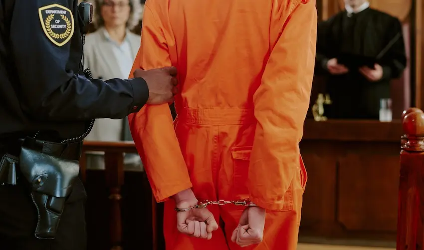 A person in an orange jail jumpsuit with hands cuffed behind their back and has a lawyer from Byron Pugh Legal is escorted by a security officer inside a courtroom while a judge stands at the bench in the background portraying the gravity of facing criminal charges during a formal court proceeding in Tennessee.