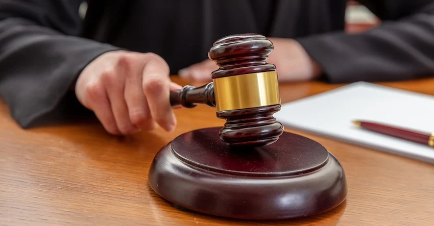 A judge wearing a black robe sits behind a warm honey-toned wooden courtroom bench and grips a dark mahogany gavel with a wide shiny gold band bringing it down firmly onto its matching oval sound block on the desk surface with their other hand resting flat on the bench beside it- a white legal notepad and a dark red ballpoint pen visible to the right of the sound block — capturing the authoritative moment a judge issues a ruling or formal decision in a criminal proceeding involving misdemeanor charges in Middle Tennessee.