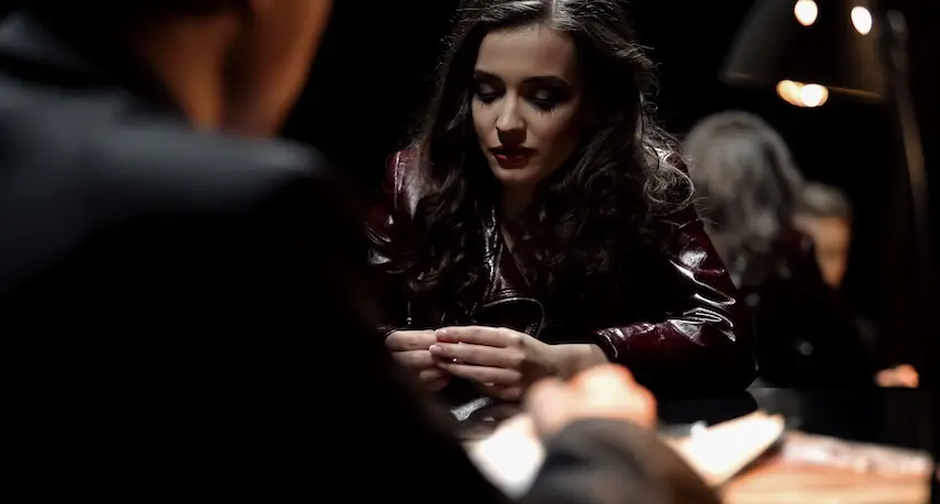 A young woman with long dark wavy hair and bold red lips wearing a deep burgundy leather jacket sits across a table from a partially blurred lawyer- from Byron Pugh Legal- in a dimly lit room illuminated by a single warm overhead lamp- her gaze cast downward toward her hands resting on the table as she listens or responds during what appears to be a tense formal legal consultation — illustrating the serious and high-stakes nature of the criminal justice process that begins when a person is facing misdemeanor charges and must navigate discussions with law enforcement or legal counsel.