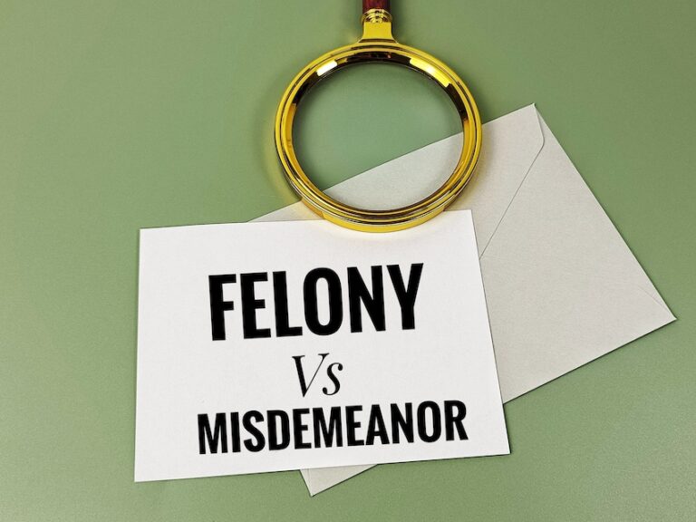 A white rectangular card printed in large bold black serif capital letters reading FELONY in the top line followed by Vs in smaller italic script in the middle- and MISDEMEANOR in large bold black capital letters across the bottom is placed on top of a light gray envelope on a muted sage green flat surface with a decorative gold-rimmed magnifying glass with a dark red wooden handle laid across the top portion of the card and envelope — a clean and direct visual comparison of the two primary tiers of criminal offenses from misdemeanor charges at the lower end to felony charges carrying the most severe legal consequences in Middle Tennessee.